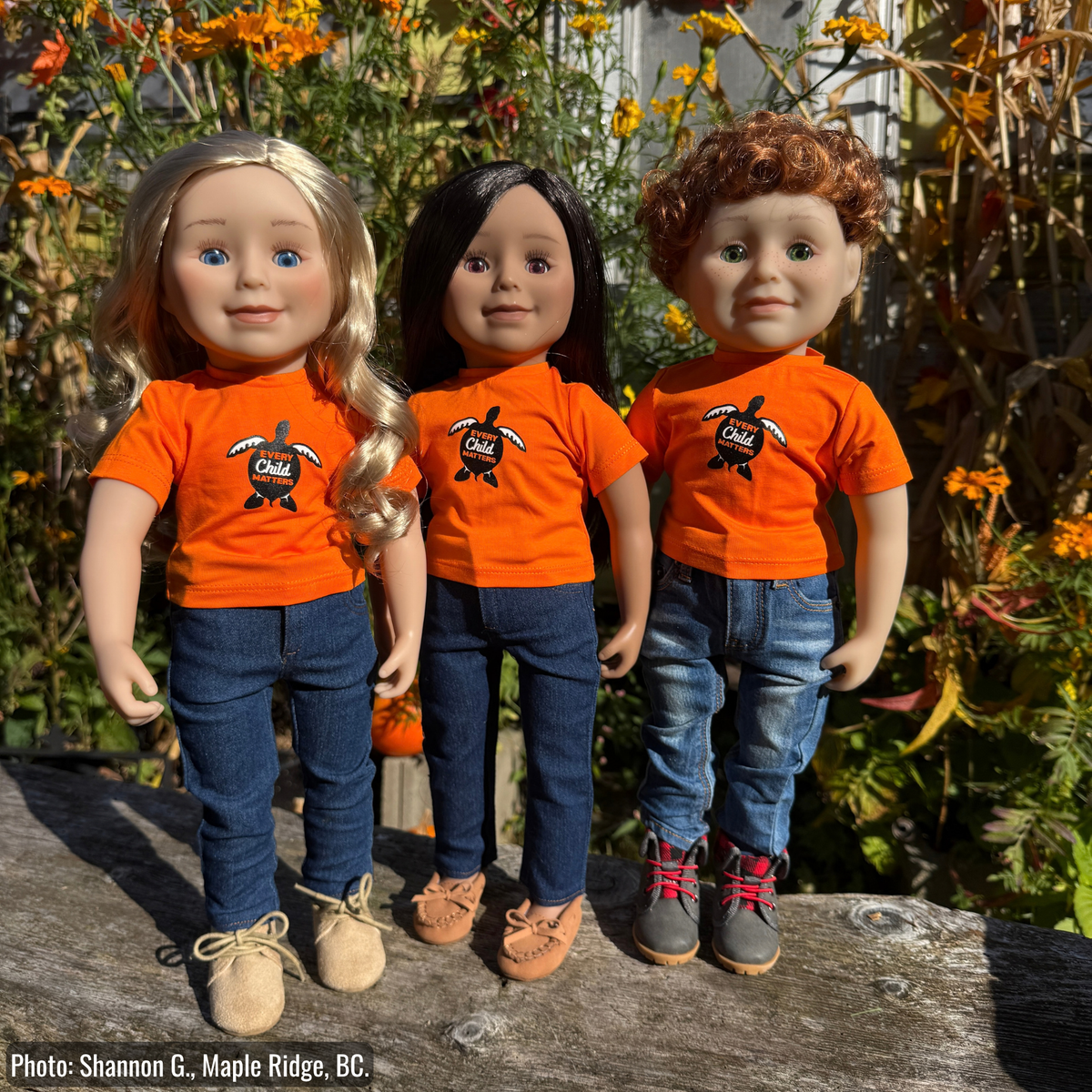 Three dolls standing outdoors on a wooden surface, wearing matching bright orange T-shirts with a turtle graphic and blue jeans.