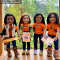 Four dolls standing together on a wooden table in front of a window with a forest view. They are wearing orange shirts, along with jeans, boots, and winter accessories, and are holding printed flyers.
