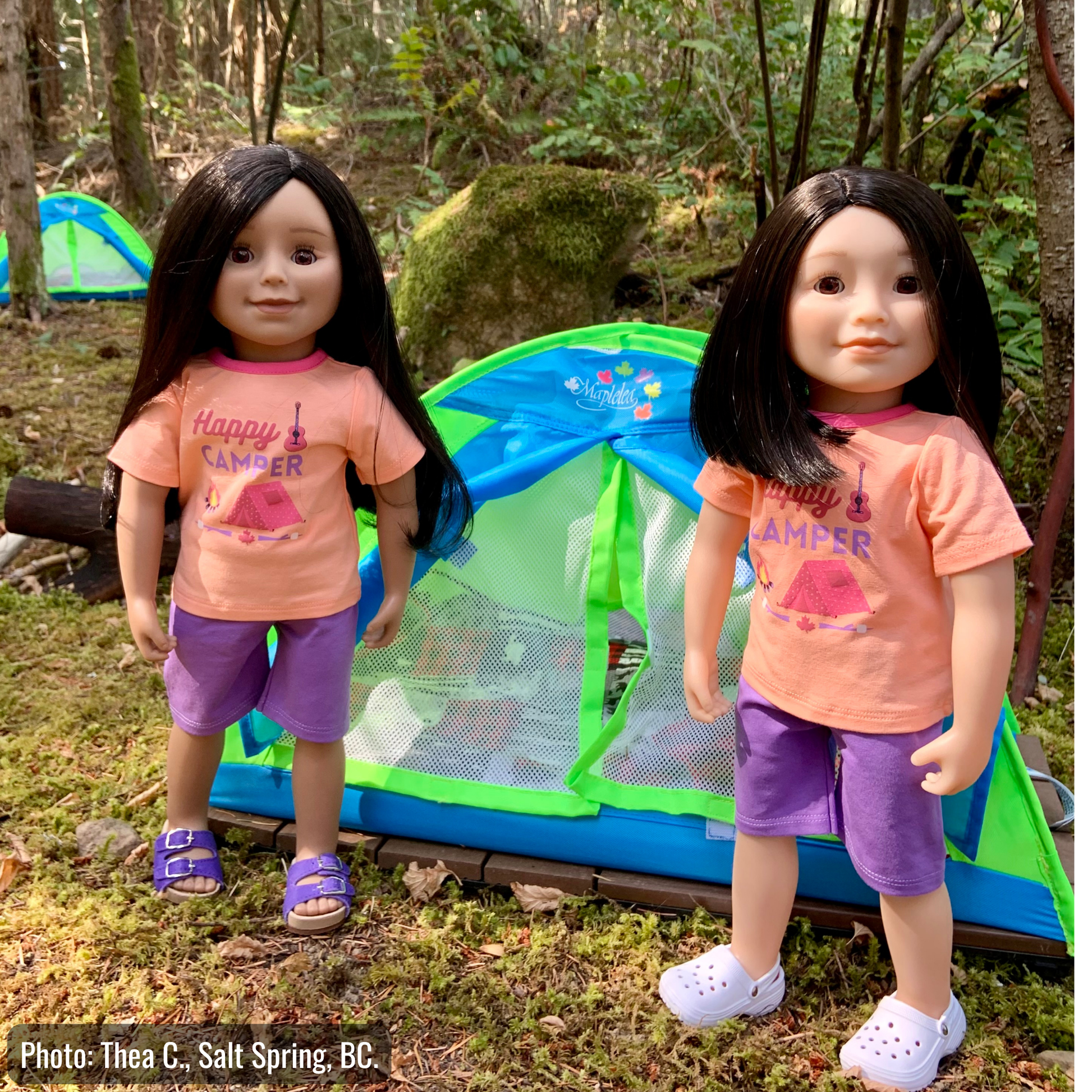 Two doll figures with long and short black hair stand in front of a small green and blue camping tent on a forest floor. Both dolls wear peach T-shirts with a pink campfire graphic and the words 'Happy Camper,' paired with purple shorts.