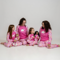 A family of four wearing matching pink long-sleeve pajamas with a white bunny graphic and floral-patterned legs, sitting together on a white studio floor.