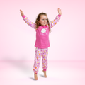 A young girl with curly brown hair is jumping barefoot with her arms raised, wearing a pink pajama set with a floral pattern. The set includes a long-sleeve top with a white bunny graphic and the text 'hope is a rabbit' on the chest, and matching patterned pants.