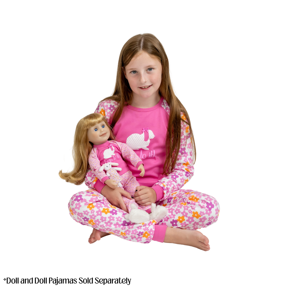 A young girl with long light brown hair sits cross-legged on a white background, wearing a pink long-sleeve pajama top with a white bunny graphic and matching floral pajama pants. She is holding a blonde doll dressed in matching pajamas.