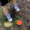 18-inch doll wearing sneakers with colorful decorations standing on a forest floor with leaves and a small orange mushroom.