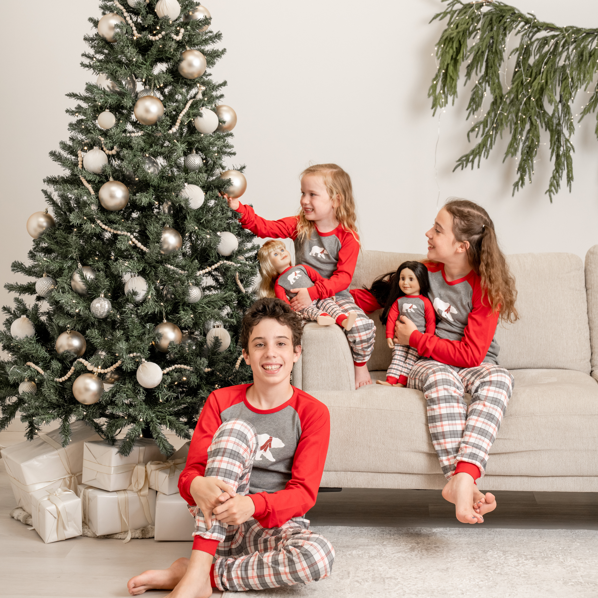 Children in matching polar bear plaid pajamas sitting on a couch with 18-inch dolls next to a decorated Christmas tree.