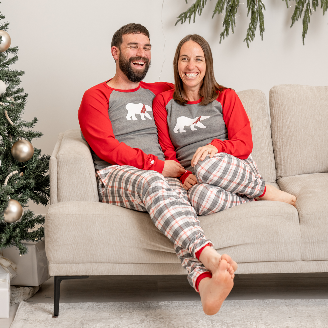 Family in matching plaid polar bear pajamas sitting on a couch with a decorated Christmas tree in the background.
