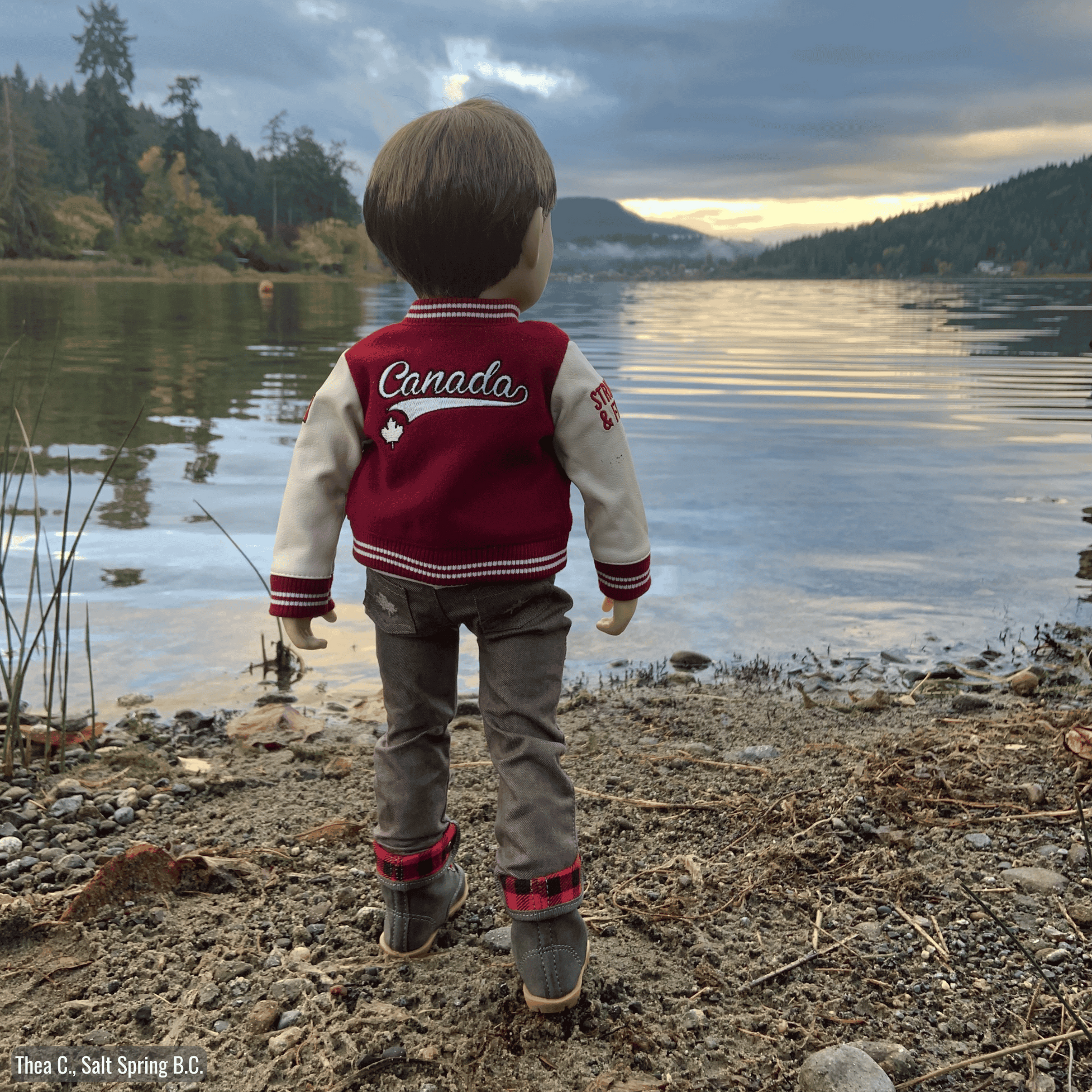 18-inch doll in a red and white varsity jacket standing by a lake with mountains in the background.