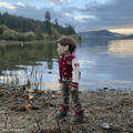 18-inch doll in a red and white varsity jacket standing by a lake with mountains in the background.