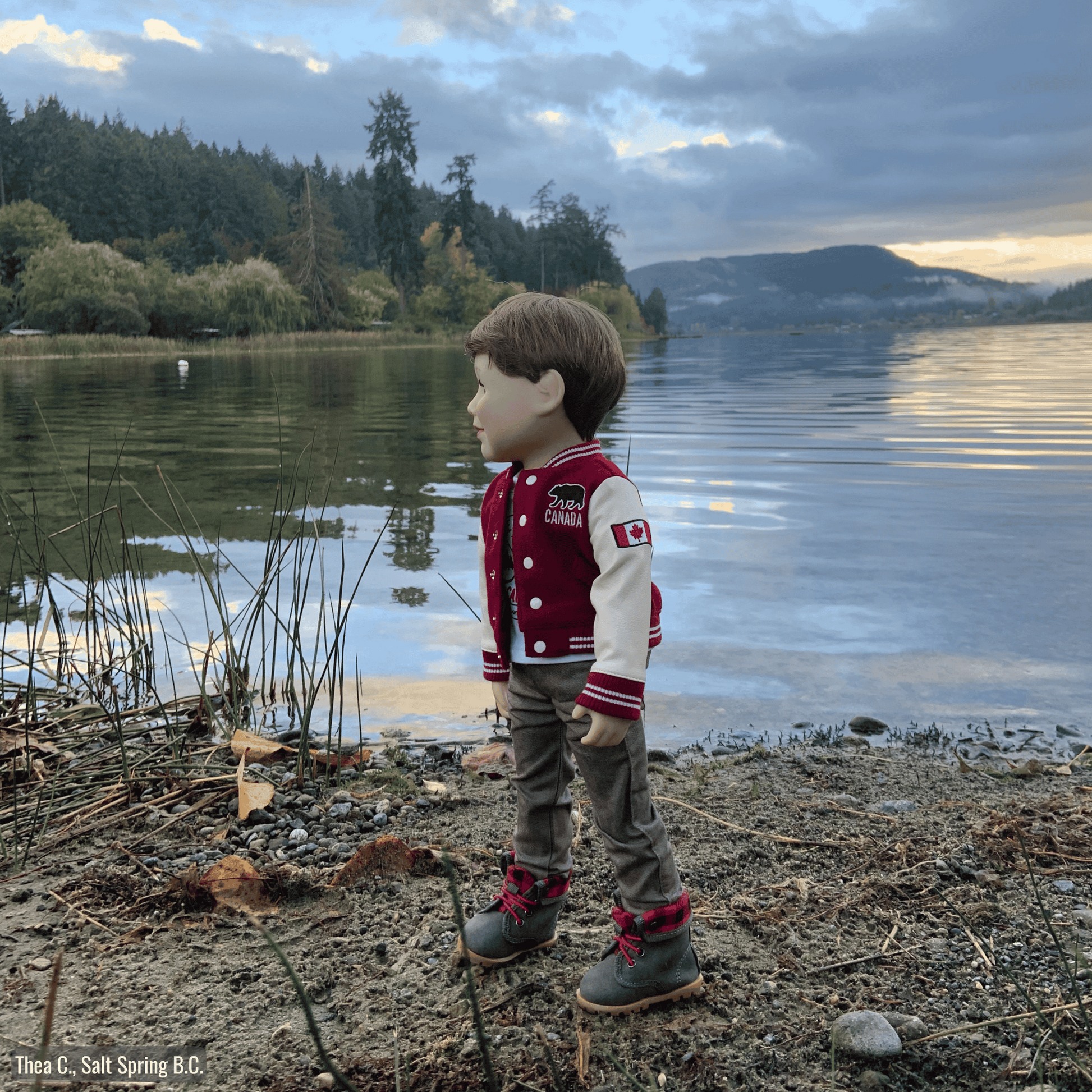 18-inch doll in a red and white varsity jacket standing by a lake with mountains in the background.