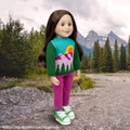 A doll with long brown hair stands outdoors in front of mountains and trees, wearing a colorful knit sweater with green sleeves and a mountain-and-sun graphic, bright pink pants, and green-and-white sneakers. Text at the bottom reads 'Flutter Field Runners Sold Separately. Taryn Maplelea doll outdoor jumper knit sweater for 18 inch dolls, 