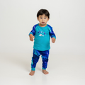 A toddler standing barefoot on a seamless white studio background, wearing a matching long-sleeve top and pants set in bright turquoise and blue with a galaxy/cityscape graphic on the chest.