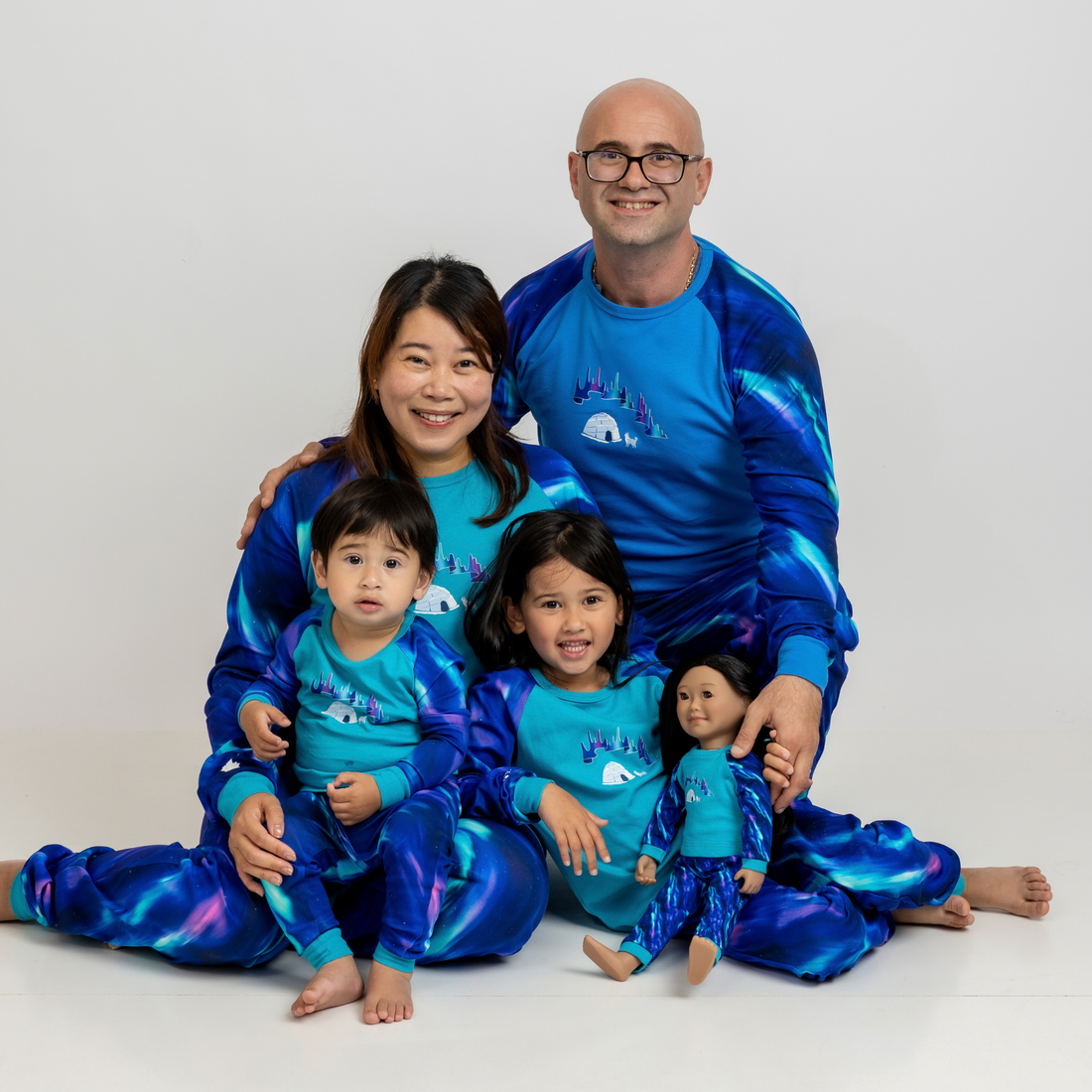 A family of four wearing matching long-sleeve pajamas with an aurora pattern. Doll wears pajamas that match the family PJs.