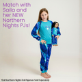 A young girl stands barefoot against a light studio background wearing a blue and turquoise long-sleeve pajama set with a matching patterned pair. She is holding a doll wearing the matching pajamas.
