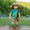 A doll dressed in outdoor camping attire stands on a dirt path in a grassy park with trees and tents in the background. The outfit includes a beige wide-brim hat with a green brim, a bright blue T-shirt with a green dinosaur graphic, tan shorts, blue socks, brown boots, and a brown crossbody bag with a neon green strap.