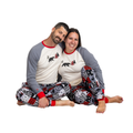A man and a woman sitting together on a white background, wearing matching long-sleeve pajamas with gray sleeves, cream bodies, red trim, and a black bear graphic on the chest, paired with patterned pants featuring red, black, white, and gray designs.