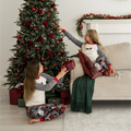 Two children wearing matching holiday pajamas decorate a Christmas tree and sit on a couch in a living room. The pajamas are white raglan-style tops with gray sleeves and patterned pants featuring red, green, and gray designs.