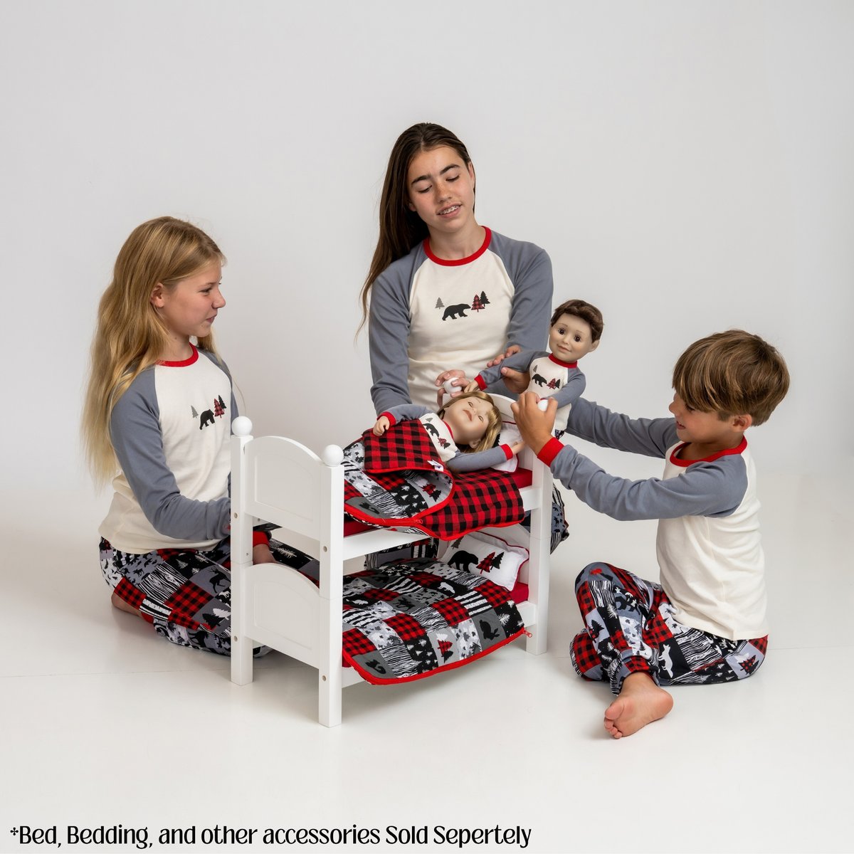 Four children wearing matching holiday pajamas play with a small white bed and plaid bedding set in a bright studio setting. The pajamas are cream and gray raglan with red trim, and the pants have a red, black, and gray plaid and animal print pattern.