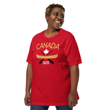 Smiling adult model wearing a red “Canada” graphic T-shirt and blue jeans against a light grey background. The shirt features the word “CANADA” in gold above a white maple leaf, a gold canoe, and crossed black paddles, with the phrase “TRUE NORTH STRONG & FREE” below. The model is shown at a slight angle looking to the side, highlighting the front design and fit of the T-shirt.