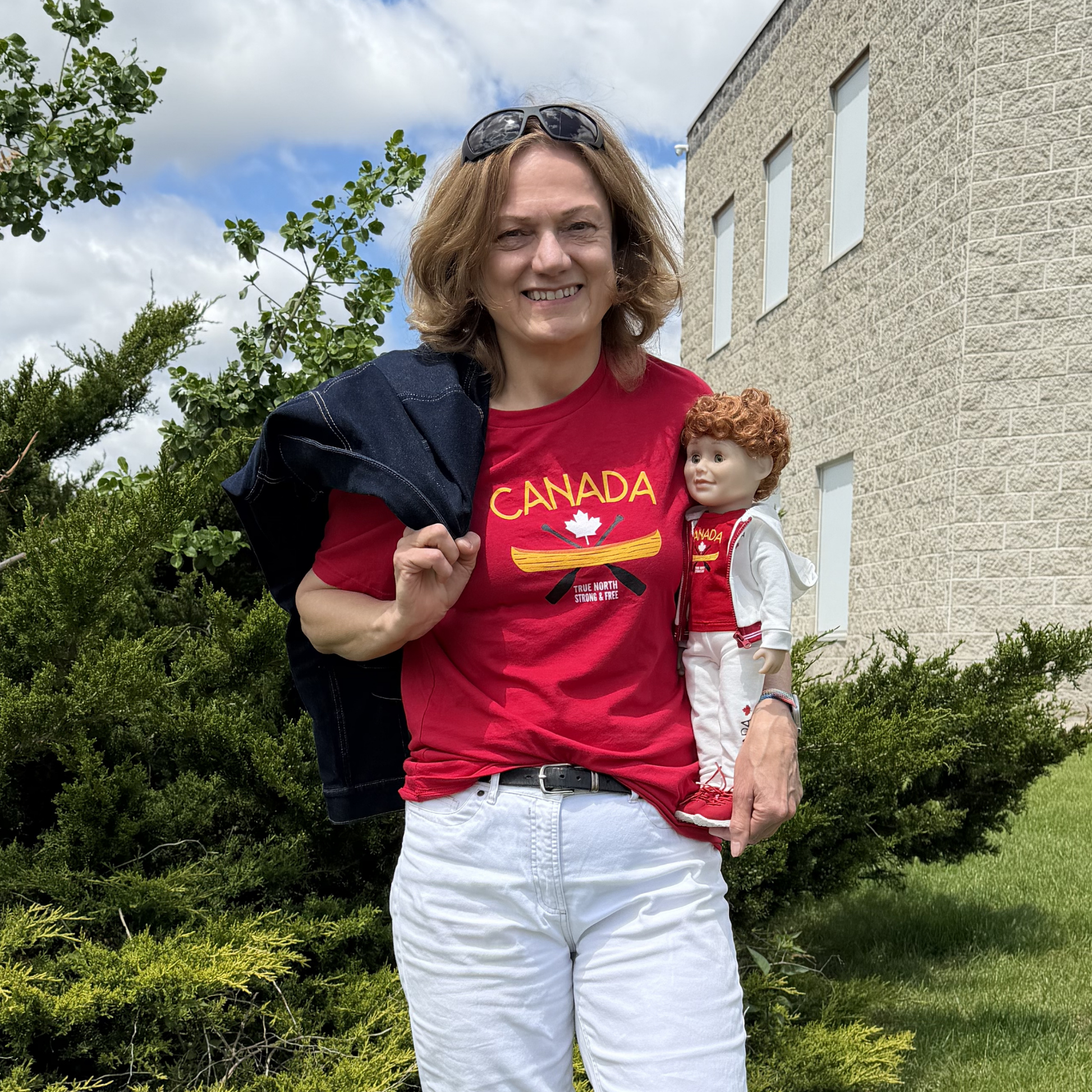 Person standing outdoors on a grassy area beside shrubs and a building, wearing a red Canada-themed graphic T-shirt and white pants. The shirt features the word “CANADA,” a white maple leaf, a gold canoe, crossed paddles, and the phrase “True North Strong & Free.” The person holds a doll dressed in a coordinating red Canada-themed outfit and carries a dark jacket over one shoulder. A partly cloudy sky is visible in the background.