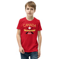 Youth model wearing a red “Canada” graphic T-shirt and blue jeans against a light grey background. The shirt features the word “CANADA” in gold above a white maple leaf, a gold canoe, and crossed black paddles, with the phrase “TRUE NORTH STRONG & FREE” below. The model faces forward with a neutral expression, showcasing the front design of the youth T-shirt.