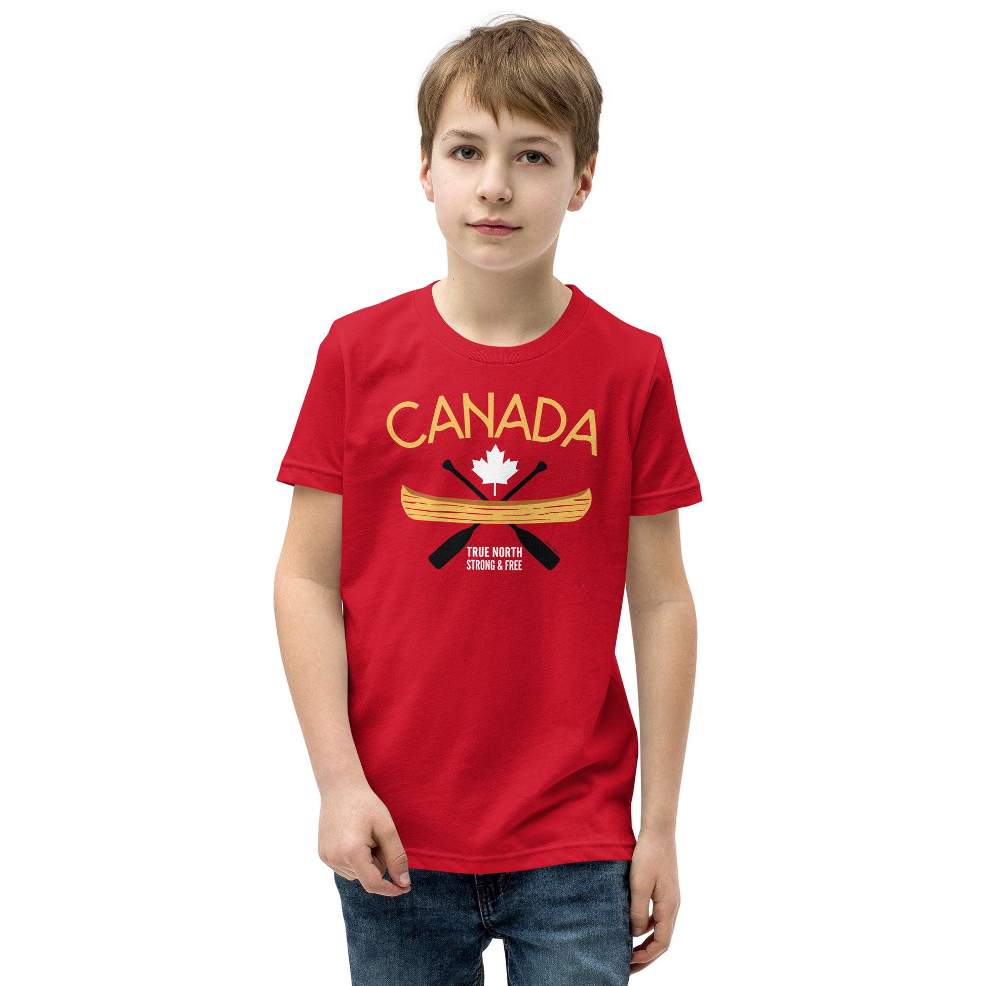 Youth model wearing a red “Canada” graphic T-shirt and blue jeans against a light grey background. The shirt features the word “CANADA” in gold above a white maple leaf, a gold canoe, and crossed black paddles, with the phrase “TRUE NORTH STRONG & FREE” below. The model faces forward with a neutral expression, showcasing the front design of the youth T-shirt.