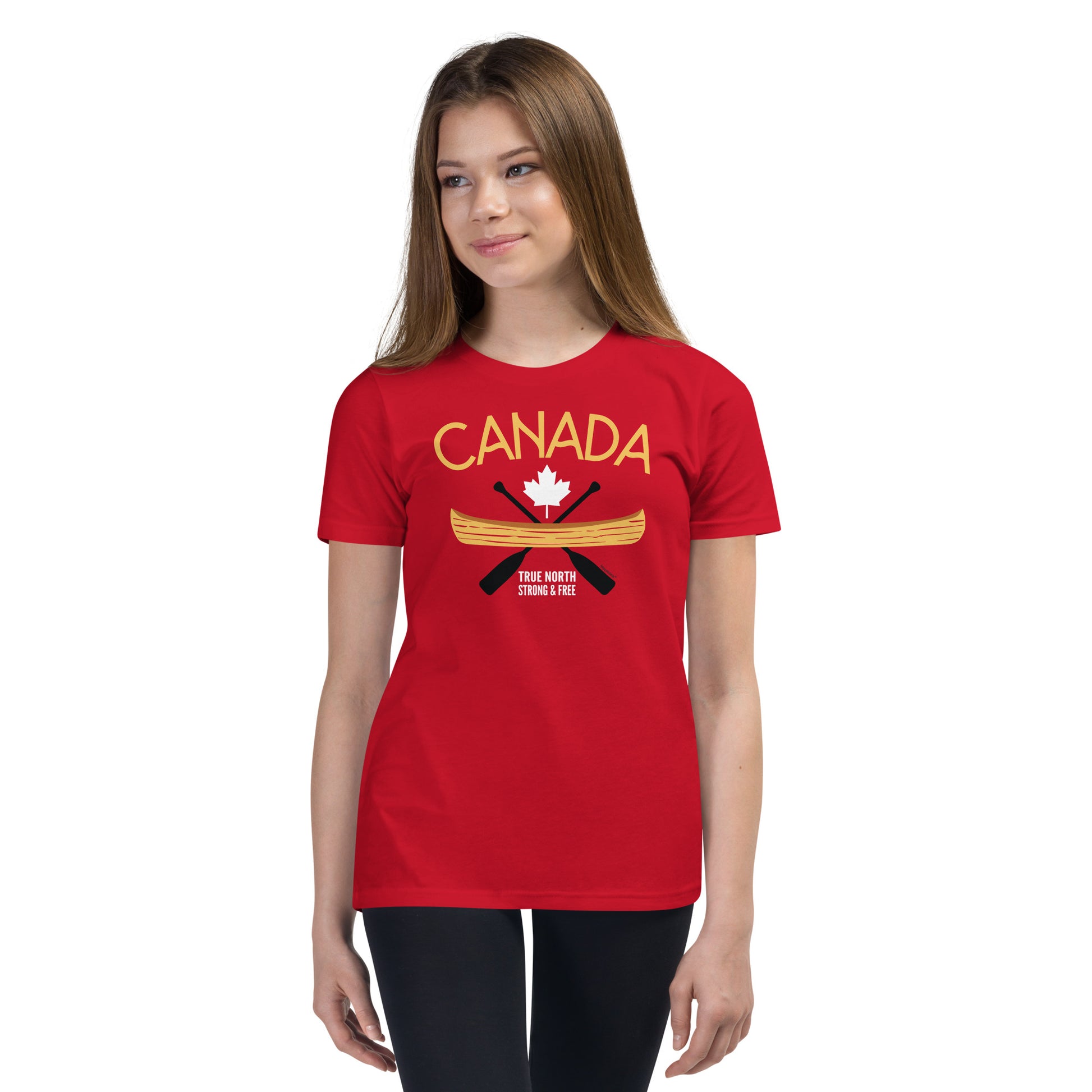 Youth model wearing a red “Canada” graphic T-shirt and dark leggings against a light grey background. The shirt features the word “CANADA” in gold above a white maple leaf, a gold canoe, and crossed black paddles, with the phrase “TRUE NORTH STRONG & FREE” below. The model is standing slightly angled and looking to the side, showcasing the front design and fit of the youth T-shirt.