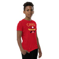 Youth model wearing a red “Canada” graphic T-shirt and black pants against a light grey background. The shirt features the word “CANADA” in gold above a white maple leaf, a gold canoe, and crossed black paddles, with the phrase “TRUE NORTH STRONG & FREE” below. The model stands at a slight angle with one hand in a pocket, showcasing the front design and side fit of the youth T-shirt.