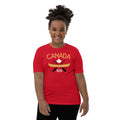 Smiling child wearing a red “Canada” graphic T-shirt against a light gray background. The shirt features a yellow canoe, crossed black paddles, a white maple leaf, and the words “Canada” and “True North Strong & Free.” The child stands facing forward, showcasing the front design of the youth T-shirt.