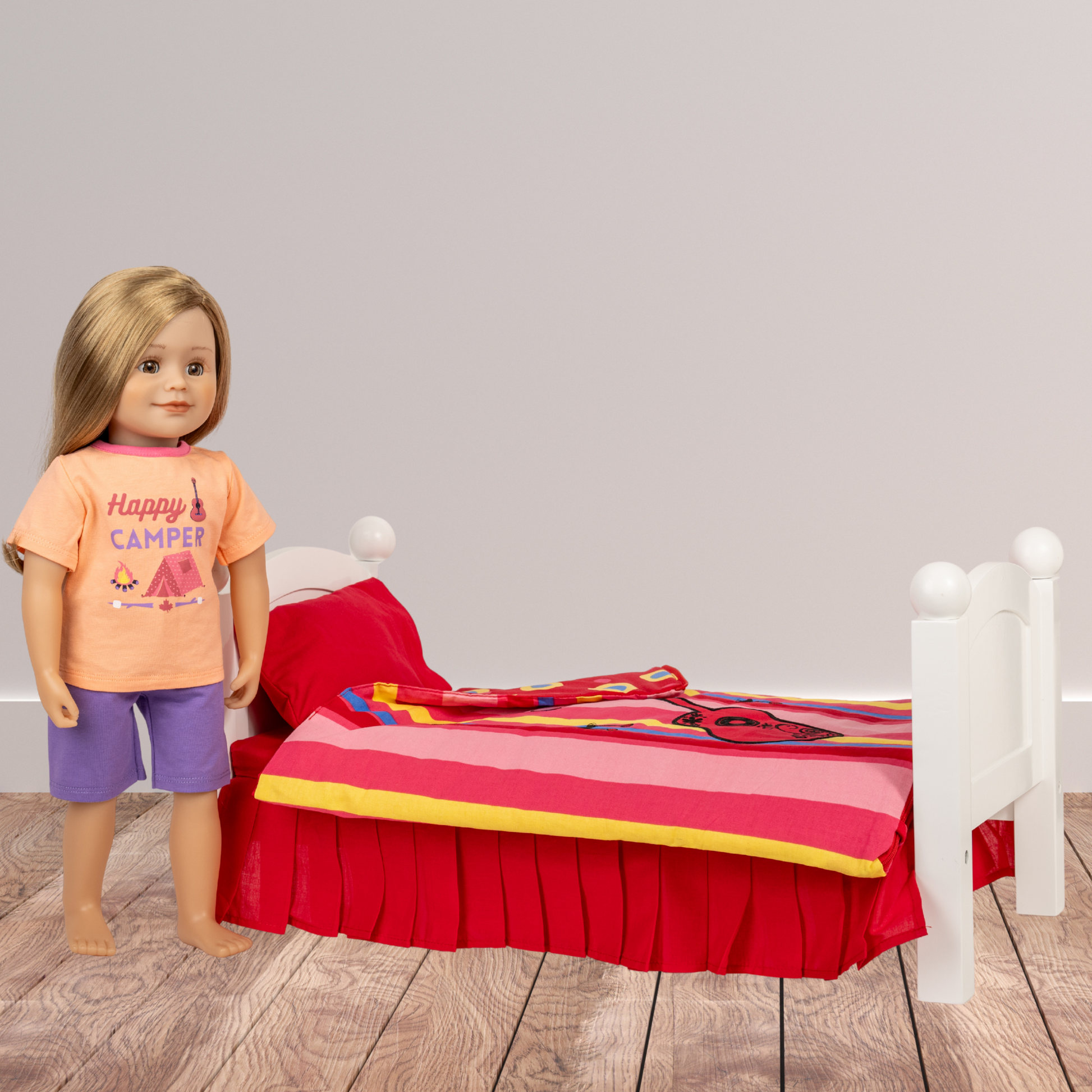 18-inch doll wearing an 'Happy Camper' shirt standing next to a bed with colourful red music themed bedding on a wooden floor.