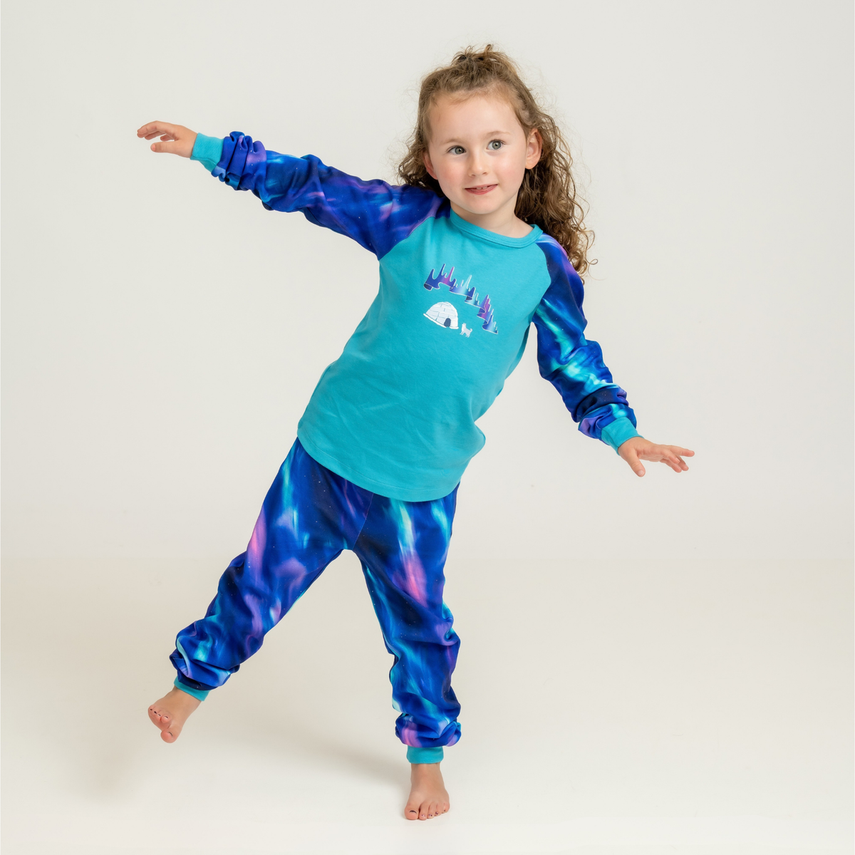 A young girl in two-piece pajamas featuring a teal and blue northern lights design with an Arctic Canada graphic, set against an aurora borealis night sky.