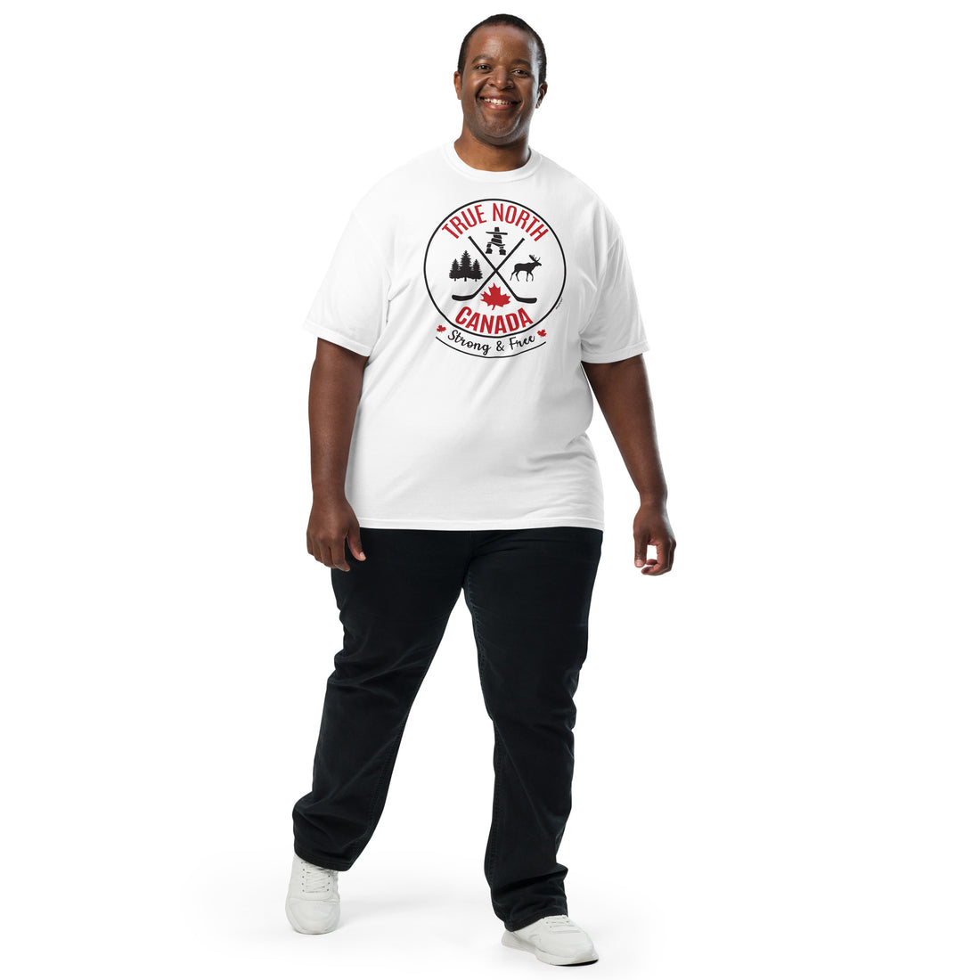 Smiling adult model wearing a white “True North Canada” graphic T-shirt, dark pants, and white sneakers, standing against a light gray background. The shirt features a circular red-and-black design with crossed hockey sticks, a maple leaf, evergreen trees, a moose, and an Inukshuk, along with the words “True North,” “Canada,” and “Strong & Free.” The model is shown walking forward, highlighting the front fit and design of the shirt.