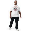 Smiling adult model wearing a white “True North Canada” graphic T-shirt, dark pants, and white sneakers, standing against a light gray background. The shirt features a circular red-and-black design with crossed hockey sticks, a maple leaf, evergreen trees, a moose, and an Inukshuk, along with the words “True North,” “Canada,” and “Strong & Free.” The model is shown walking forward, highlighting the front fit and design of the shirt.