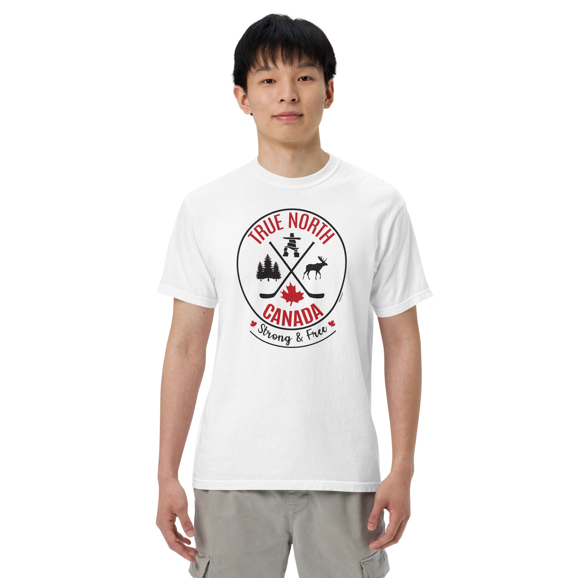 Adult model wearing a white “True North Canada” graphic T-shirt and light gray pants, standing against a light gray background. The shirt features a circular red-and-black design with crossed hockey sticks, a maple leaf, evergreen trees, a moose, and an Inukshuk, along with the words “True North,” “Canada,” and “Strong & Free.” The model faces forward with a neutral expression, showcasing the front of the shirt.