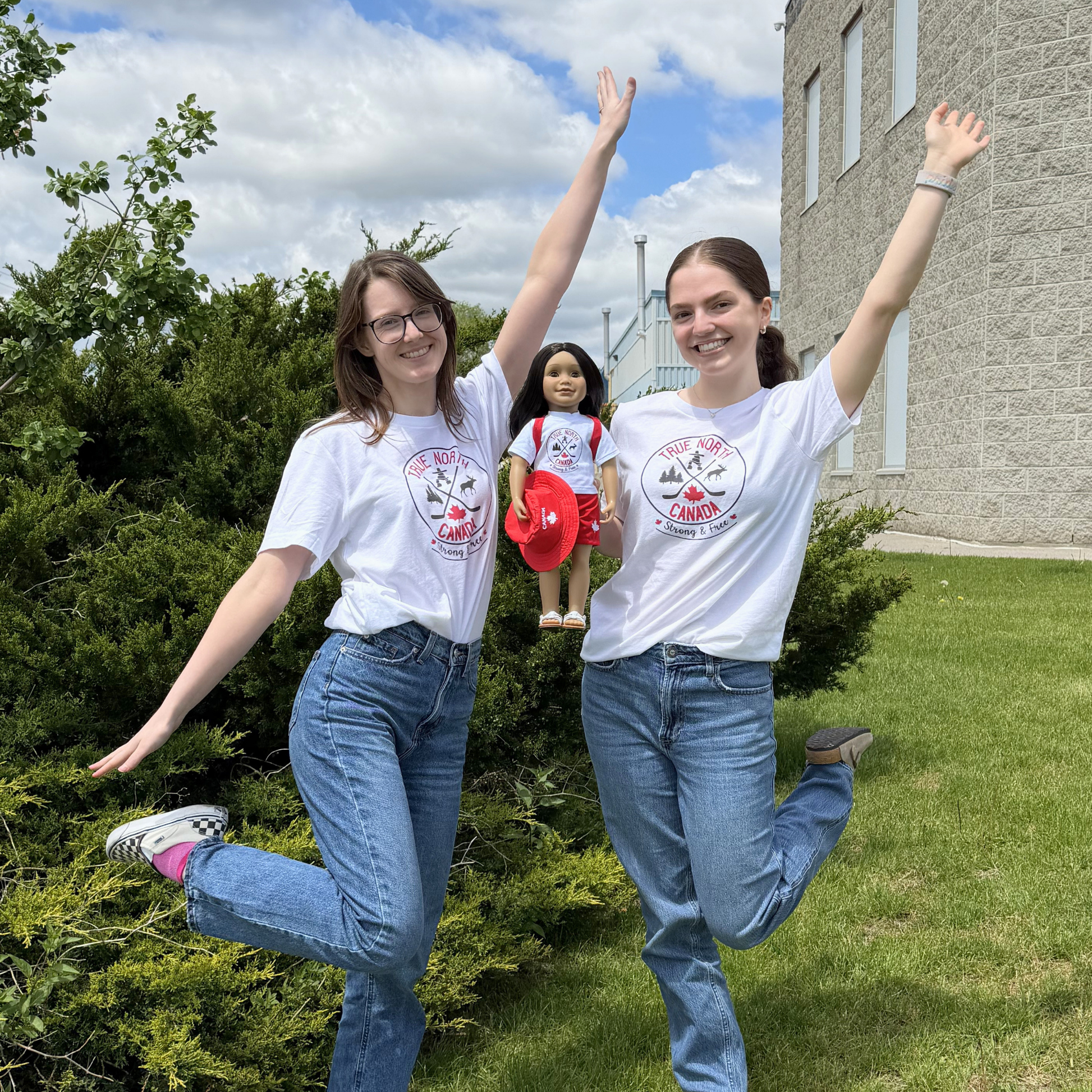 Two young adults stand outdoors on a grassy area beside shrubs and a building, smiling with their arms raised. Both are wearing white “True North Canada” graphic T-shirts and blue jeans. Between them is a doll dressed in a matching “True North Canada” T-shirt, red shorts, and a red Canada-themed hat. The scene is set under a partly cloudy sky, highlighting the matching outfits for adults and dolls.
