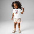 A young child with curly dark hair stands against a light gray background wearing a white T-shirt featuring a circular “True North Canada” graphic with crossed hockey sticks, a maple leaf, and wildlife silhouettes. The child also wears light pink heart-patterned shorts, matching socks, and beige sneakers, looking down with one hand raised near their hair.