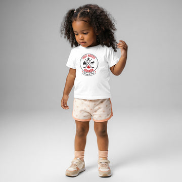 A young child with curly dark hair stands against a light gray background wearing a white T-shirt featuring a circular “True North Canada” graphic with crossed hockey sticks, a maple leaf, and wildlife silhouettes. The child also wears light pink heart-patterned shorts, matching socks, and beige sneakers, looking down with one hand raised near their hair.