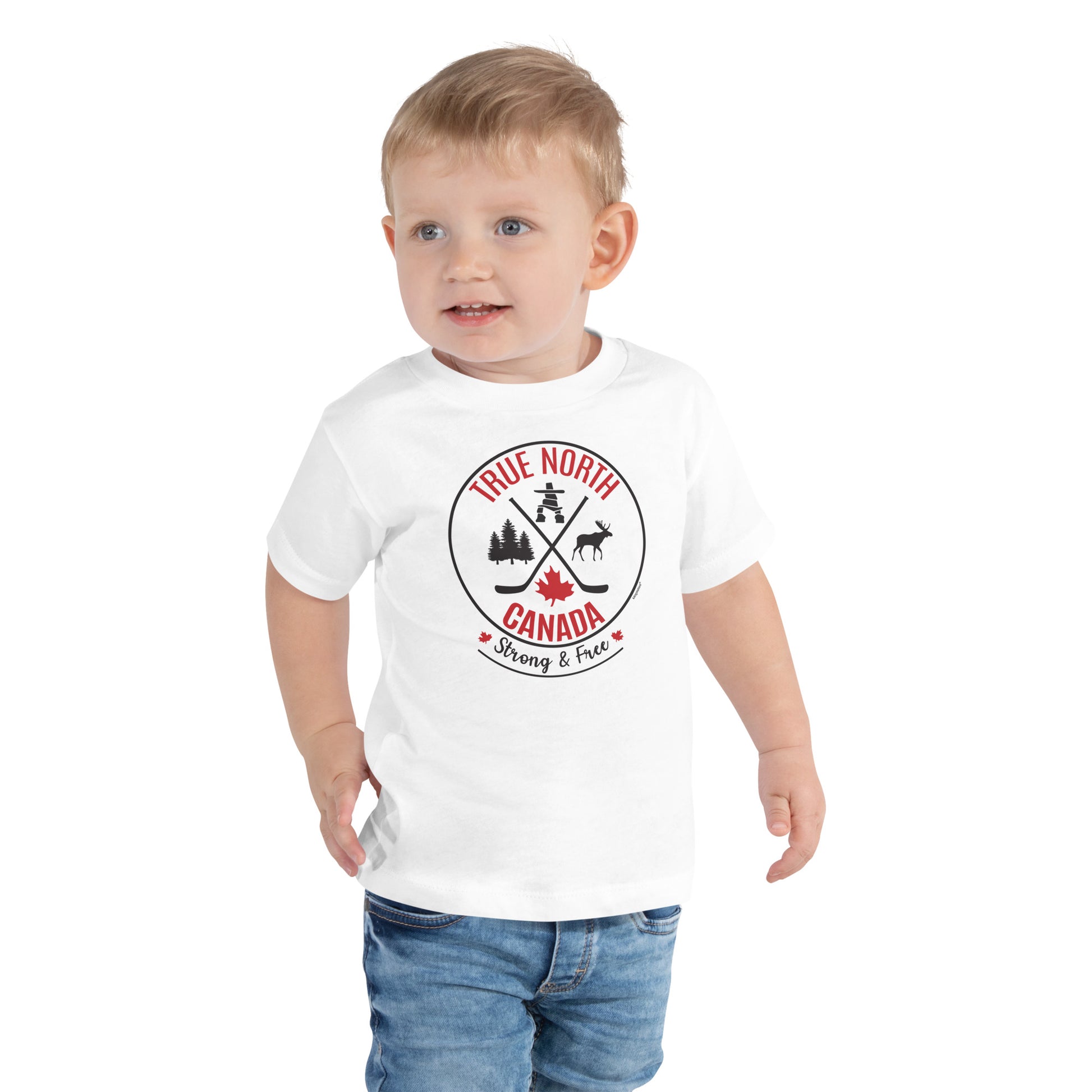 Toddler wearing a white “True North Canada” graphic T-shirt and blue jeans, standing against a light gray background. The shirt features a circular red-and-black design with crossed hockey sticks, a maple leaf, evergreen trees, a moose, and an Inukshuk, along with the words “True North,” “Canada,” and “Strong & Free.” The child is looking slightly to the side with a cheerful expression.