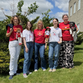 Five people stand together outdoors on a grassy lawn in front of shrubs and a building under a partly cloudy sky. They are wearing Canada-themed graphic T-shirts in red and white, with two people holding dolls dressed in coordinating Canada-themed outfits. One person on the far right holds a small black-and-white dog wearing a red bandana. The group smiles at the camera, showcasing matching apparel for adults and dolls.