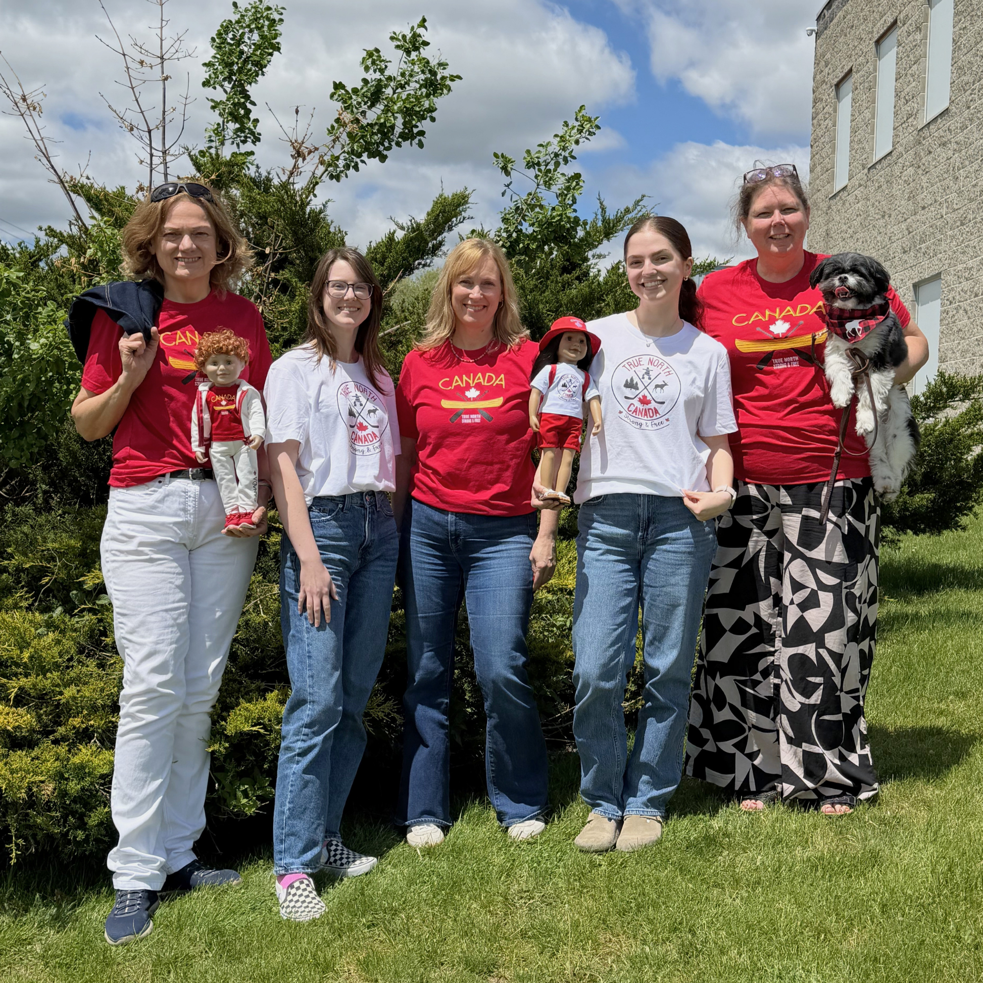 Five people stand together outdoors on a grassy lawn in front of shrubs and a building under a partly cloudy sky. They are wearing Canada-themed graphic T-shirts in red and white, with two people holding dolls dressed in coordinating Canada-themed outfits. One person on the far right holds a small black-and-white dog wearing a red bandana. The group smiles at the camera, showcasing matching apparel for adults and dolls.