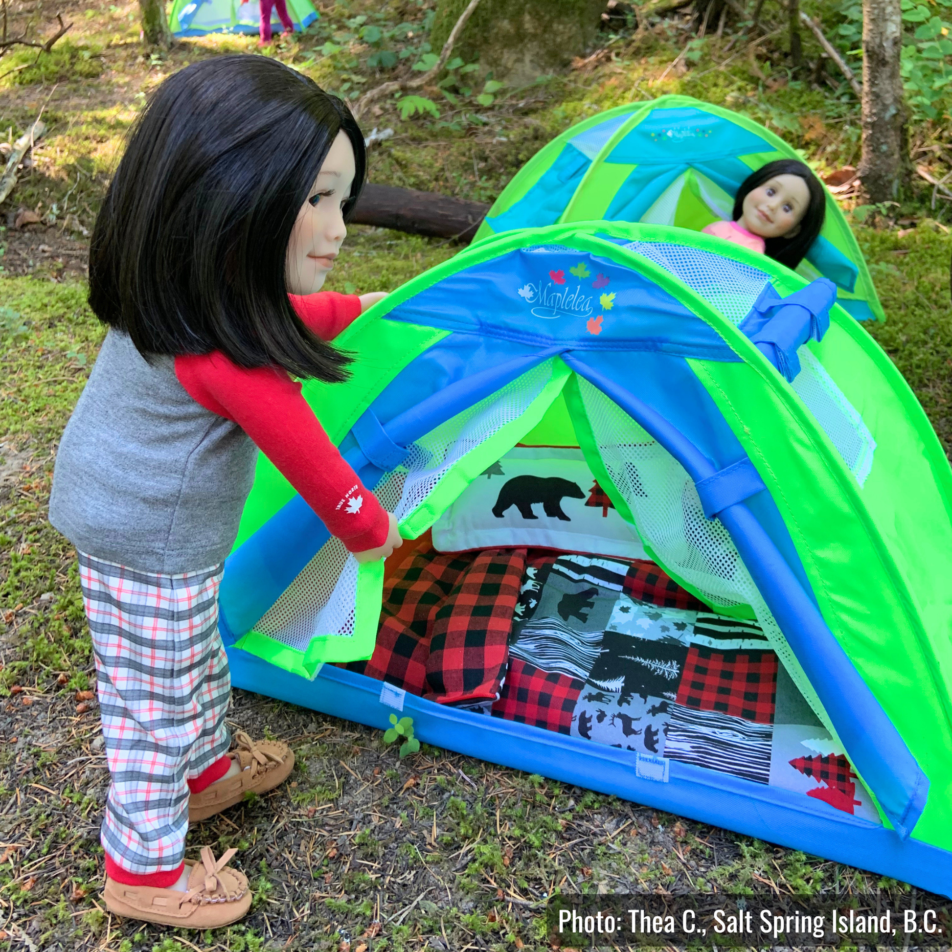 Two dolls with straight black hair are camping in a forest clearing beside bright green-and-blue toy tents. One doll, wearing a grey-and-red pyjama top, plaid pyjama pants, and moccasins, opens the flap of a tent decorated with a red-and-black buffalo plaid blanket and woodland-themed bedding, while another doll relaxes in a tent behind her. Moss-covered ground, trees, and dappled sunlight create an outdoor campsite setting.